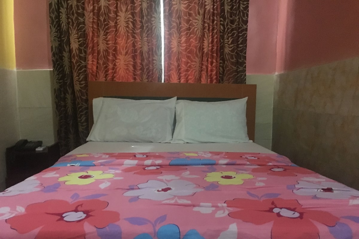 A bed with a floral bedspread, two pillows, and a wooden headboard in a room with patterned curtains and a tiled wall.