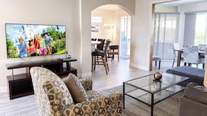 Una sala de estar moderna con televisor de pantalla plana, sofá estampado, mesa de centro de vidrio y un comedor con sillas y mesa.