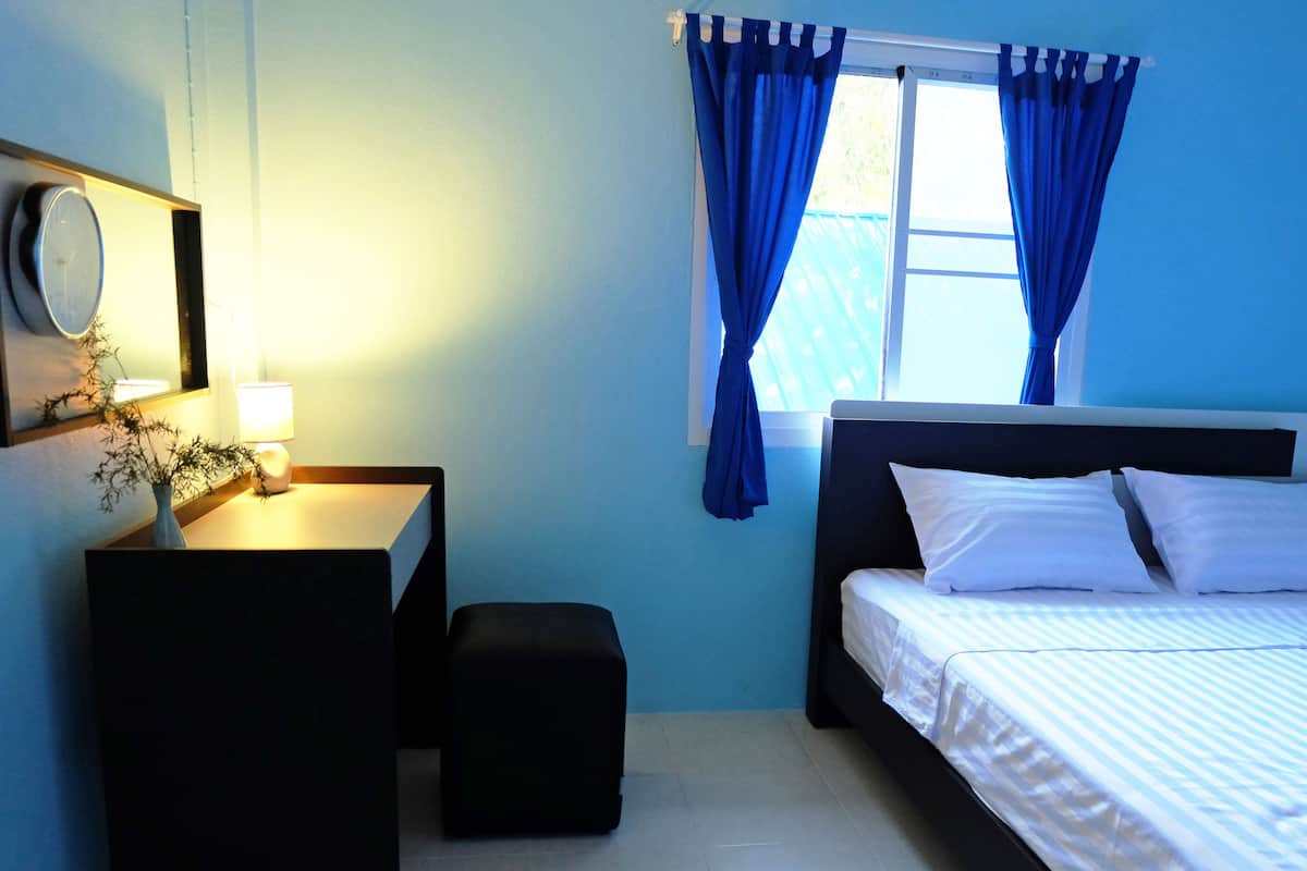A bedroom with a bed, a desk, a chair, a small table with a lamp, and a window with blue curtains.