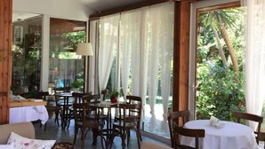 A room with wooden beams, a chandelier, a dining area with tables and chairs, and large windows with curtains.
