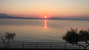 A calm lake at sunset with a clear sky, a distant mountain range, and a tree-lined path.