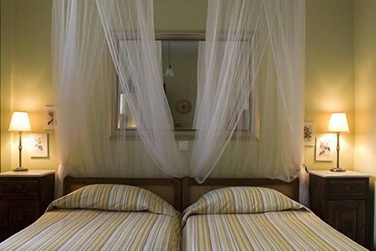 Ein Zimmer mit zwei Betten, jeweils mit gestreifter Bettwäsche und einem Baldachin. An jeder Seite der Betten befinden sich Nachttischlampen.