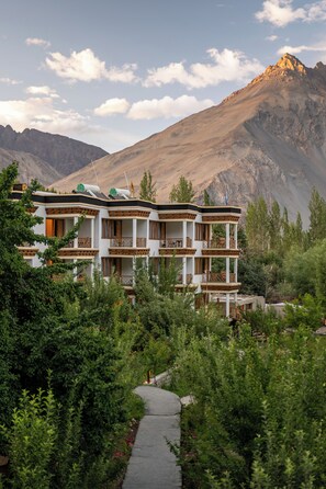 A multi-story building with balconies, surrounded by greenery and mountains in the background.