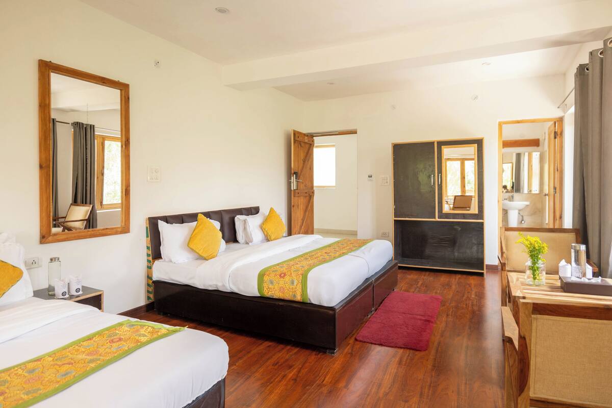 A hotel room with two beds, wooden flooring, a desk with a chair, a mirror, and a window.