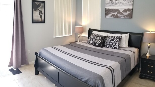 A bedroom with a bed, two bedside tables, a window with blinds, a picture on the wall, and a curtain. - Modern pool home with Game Room, 2 en suites and just 15 minutes to Theme Parks! (Davenport)