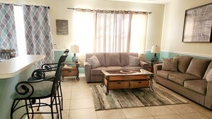 A living room with a sofa, a coffee table, a dining table with chairs, and a kitchen area. - Modern pool home with Game Room, 2 en suites and just 15 minutes to Theme Parks! (Davenport)
