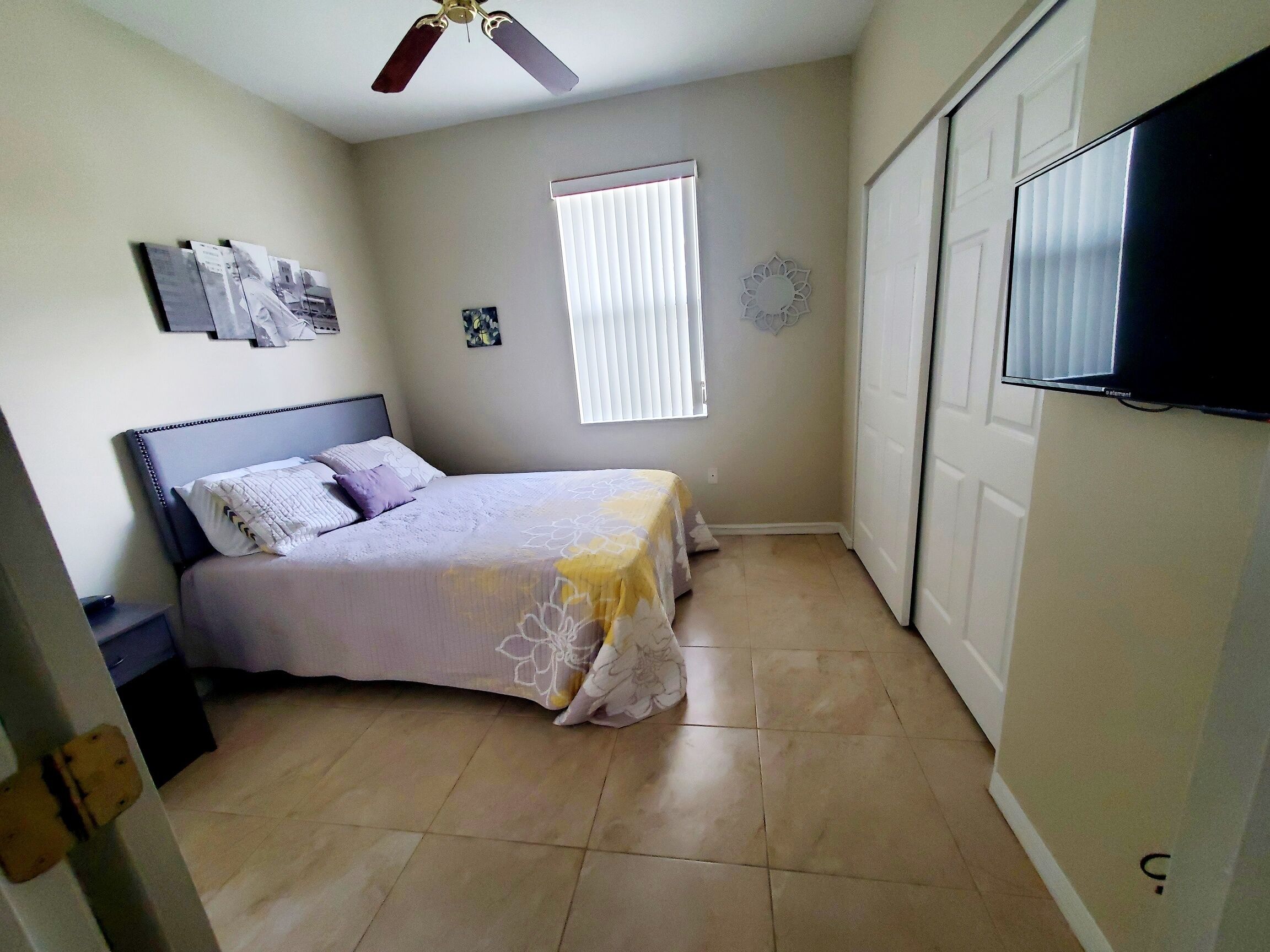 A small bedroom with a bed, a ceiling fan, a window with blinds, and a TV mounted on the wall.