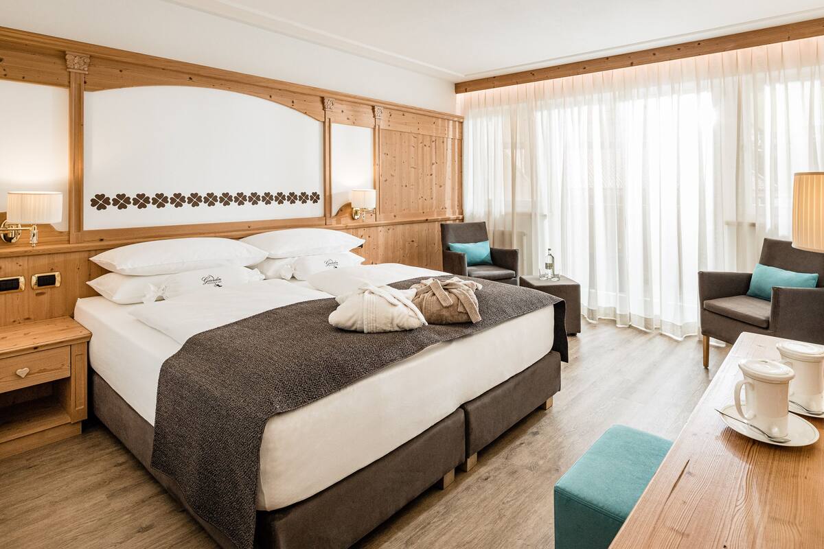 A hotel room with a large bed, a wooden headboard, a seating area with a table and chairs, and a window with sheer curtains.