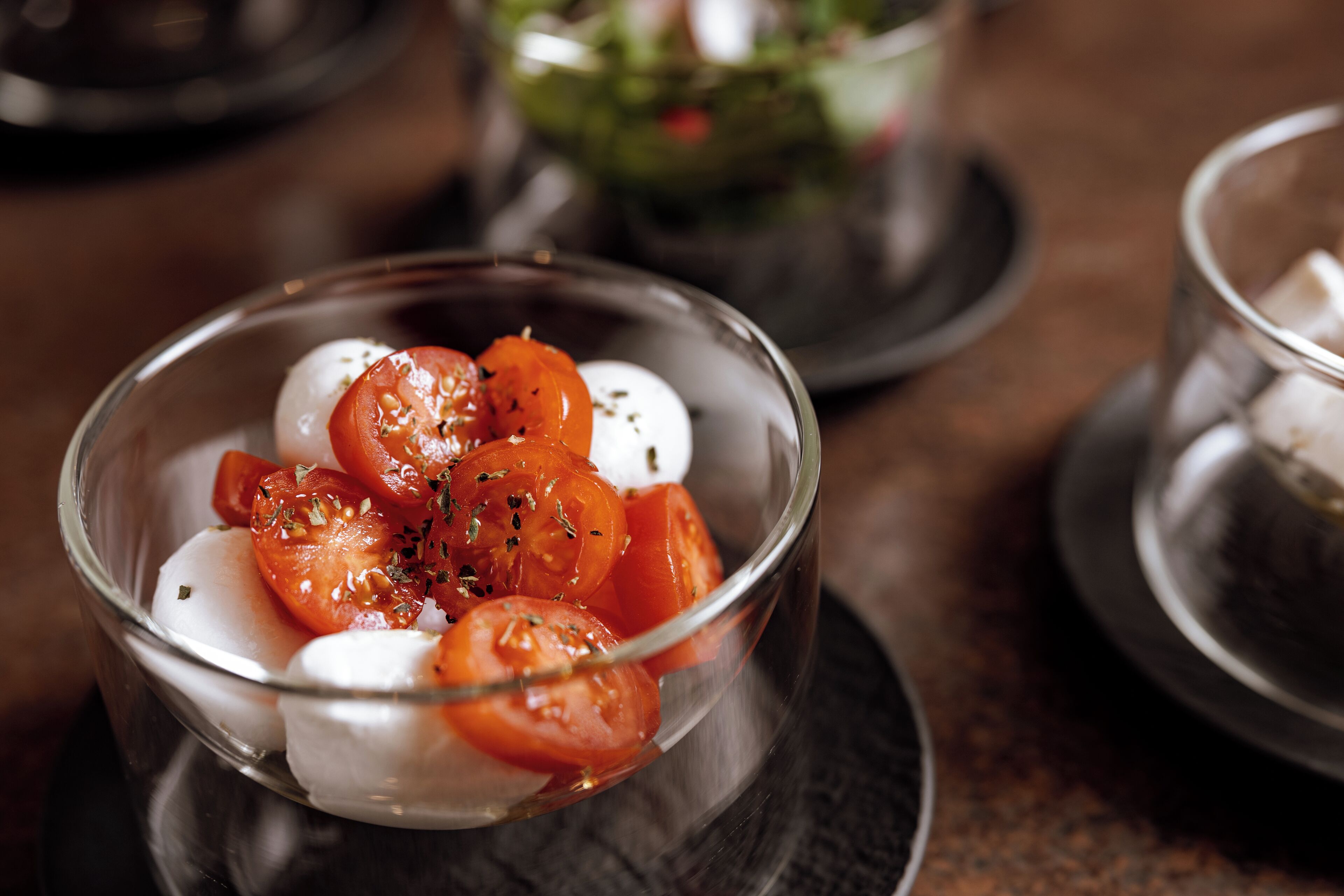 A clear glass bowl containing sliced tomatoes and mozzarella on a dark plate.
