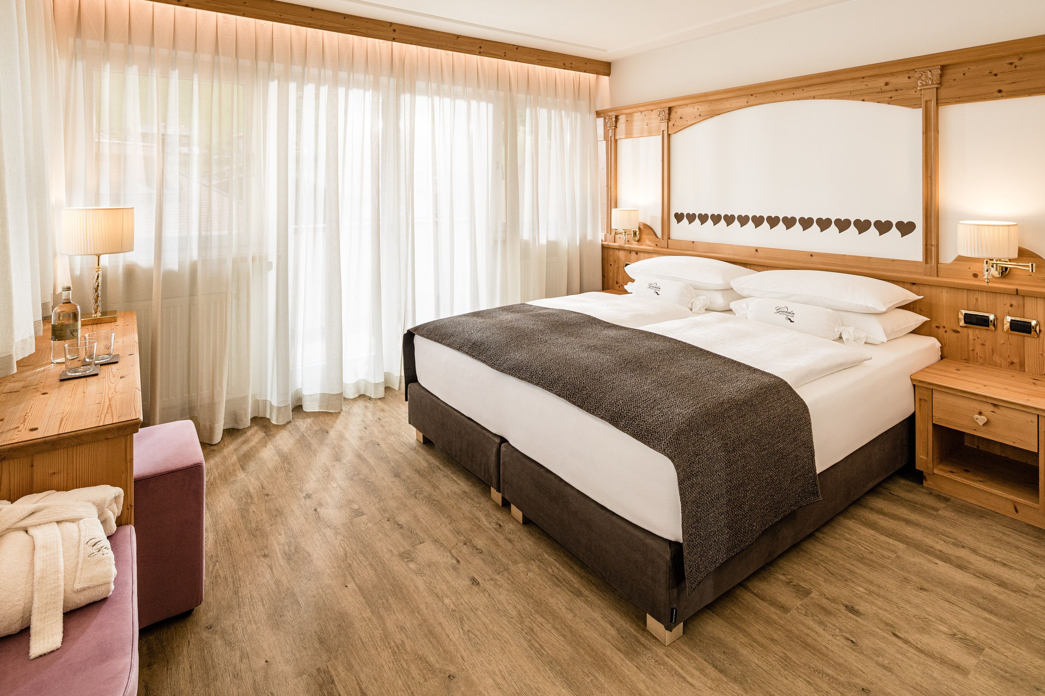 Habitación de hotel con una cama grande, cabecero de madera, mesitas de noche con lámparas, una banqueta de madera y un amplio ventanal con cortinas translúcidas.