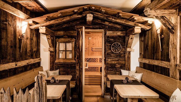 A rustic wooden cabin with wooden benches and tables, a wooden door, and a wheel in the background. - Giardin Boutique Hotel B&B (Selva di Val Gardena)
