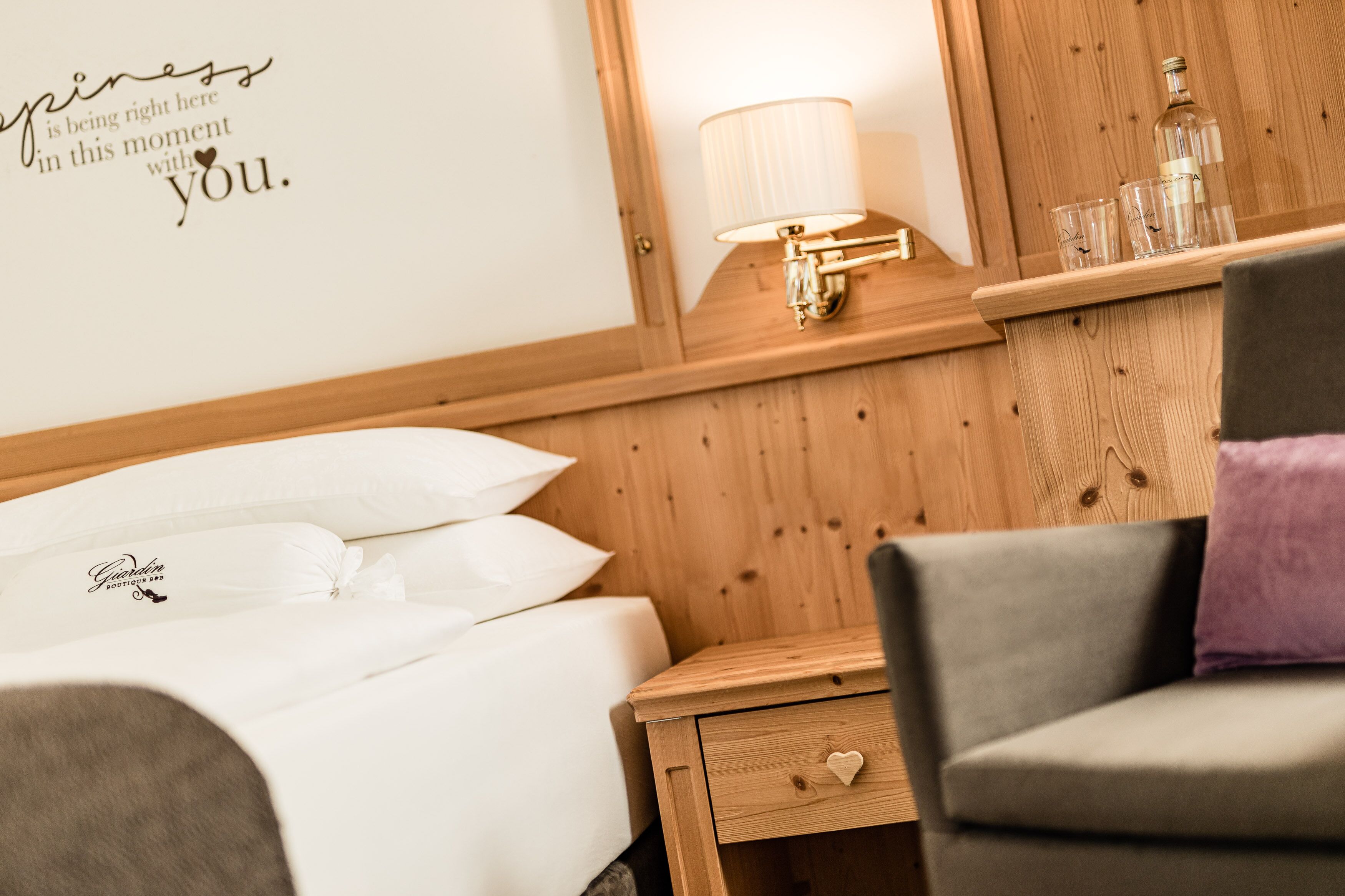 Una habitación de hotel con cabecera de madera, lámpara de noche, mesita de noche con cajón y cama con ropa blanca.