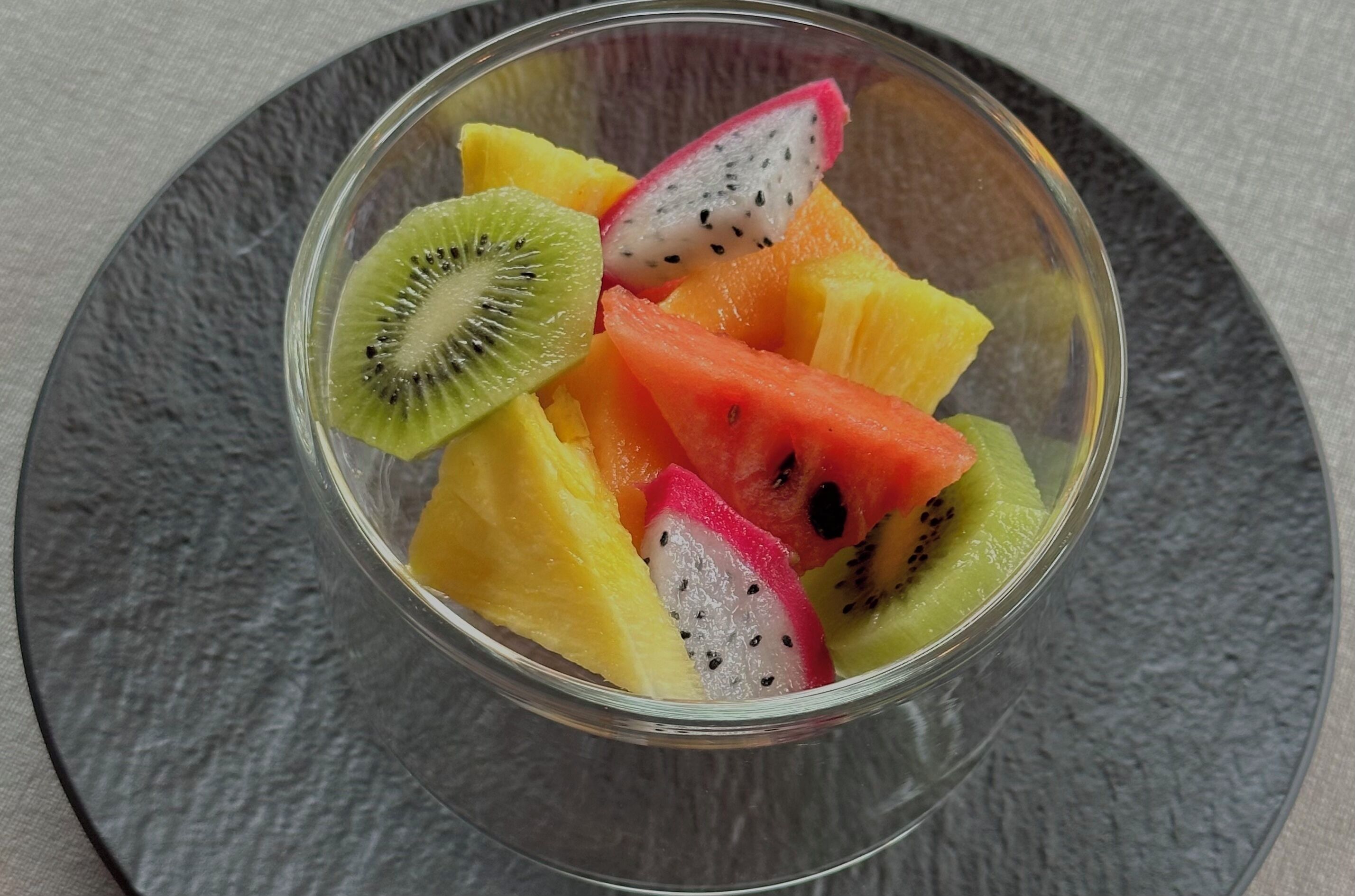 Eine transparente Glasschale mit verschiedenen Früchten, darunter Kiwis, Drachenfrüchte und Wassermelone, auf einer strukturierten, grauen Oberfläche.