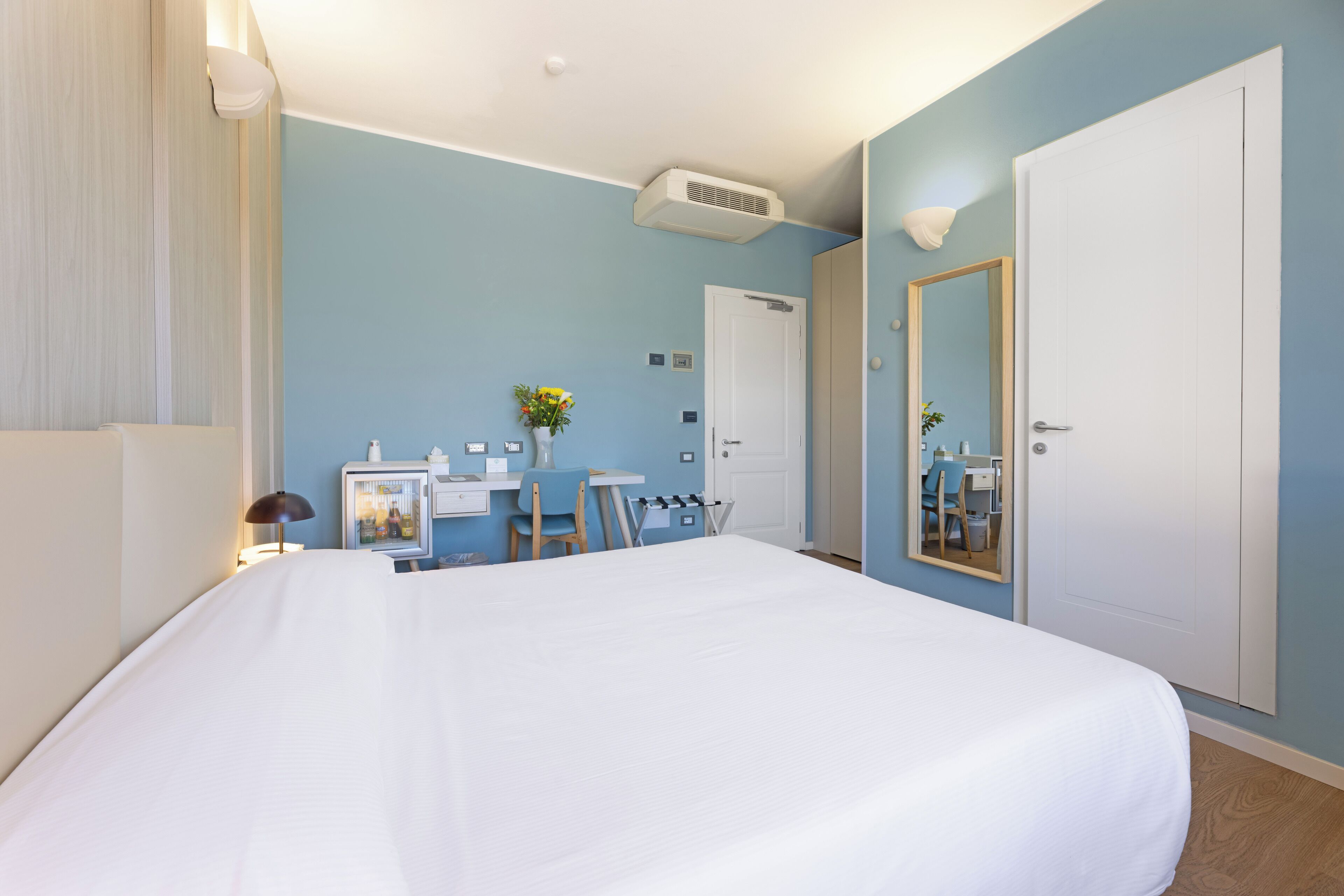 Quarto de hotel com uma cama grande, uma escrivaninha com cadeira, uma mesinha com um vaso de flores, um espelho e um guarda-roupa.