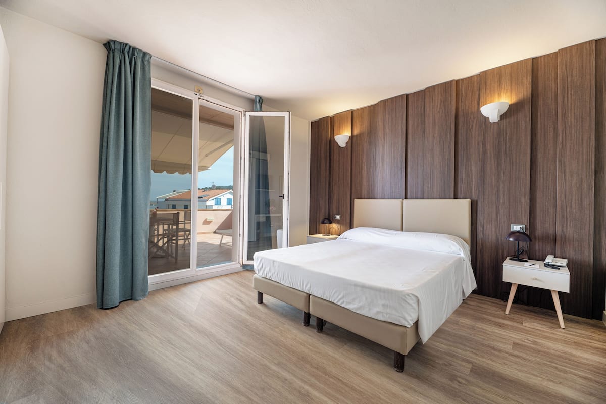A modern hotel room with a large bed, a sliding glass door to a balcony, a bedside table with a lamp, and wooden paneling on the walls.