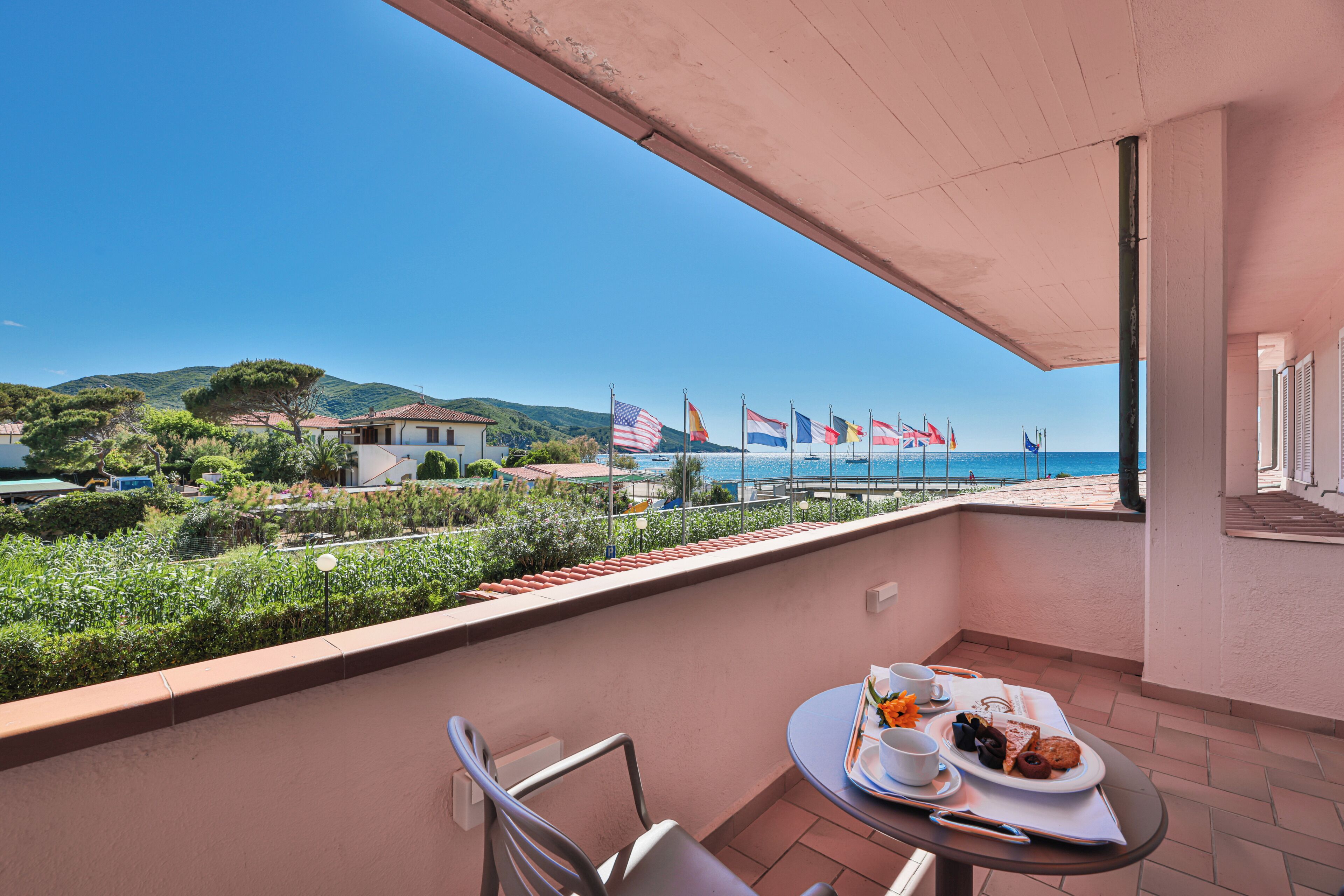 A balcony with a table set for breakfast, overlooking a coastal area with flags and a view of the sea.