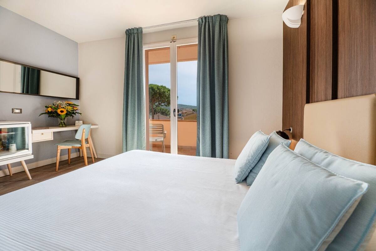 A hotel room with a large bed, a desk with a chair, a mirror, and a view of the outdoors through a sliding door.