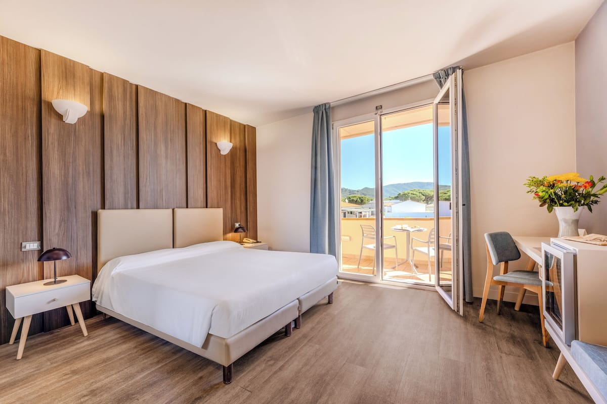 A modern hotel room with a large bed, a desk, and a balcony with a view.