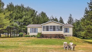 A single-story house with a covered porch, surrounded by a grassy yard and dense trees. - Finger Lakes Retreat w/ Sunroom, Fire Pit & BBQ! (Himrod)