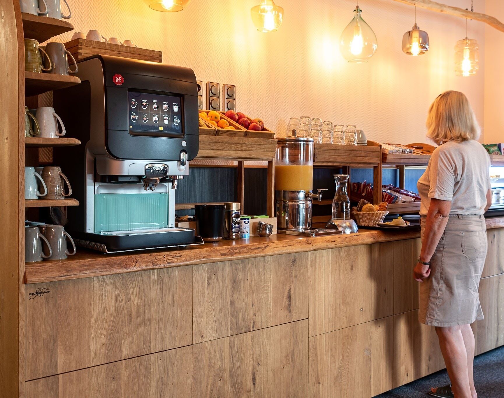 Een koffiebar met een koffieapparaat, een melkopschuimer en een koffiemolen. Er zijn planken met bekers en een fruitmand.