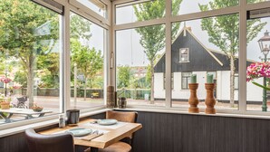 A dining area with a table and chairs, large windows with a view of the outside, and a dark-colored wall.