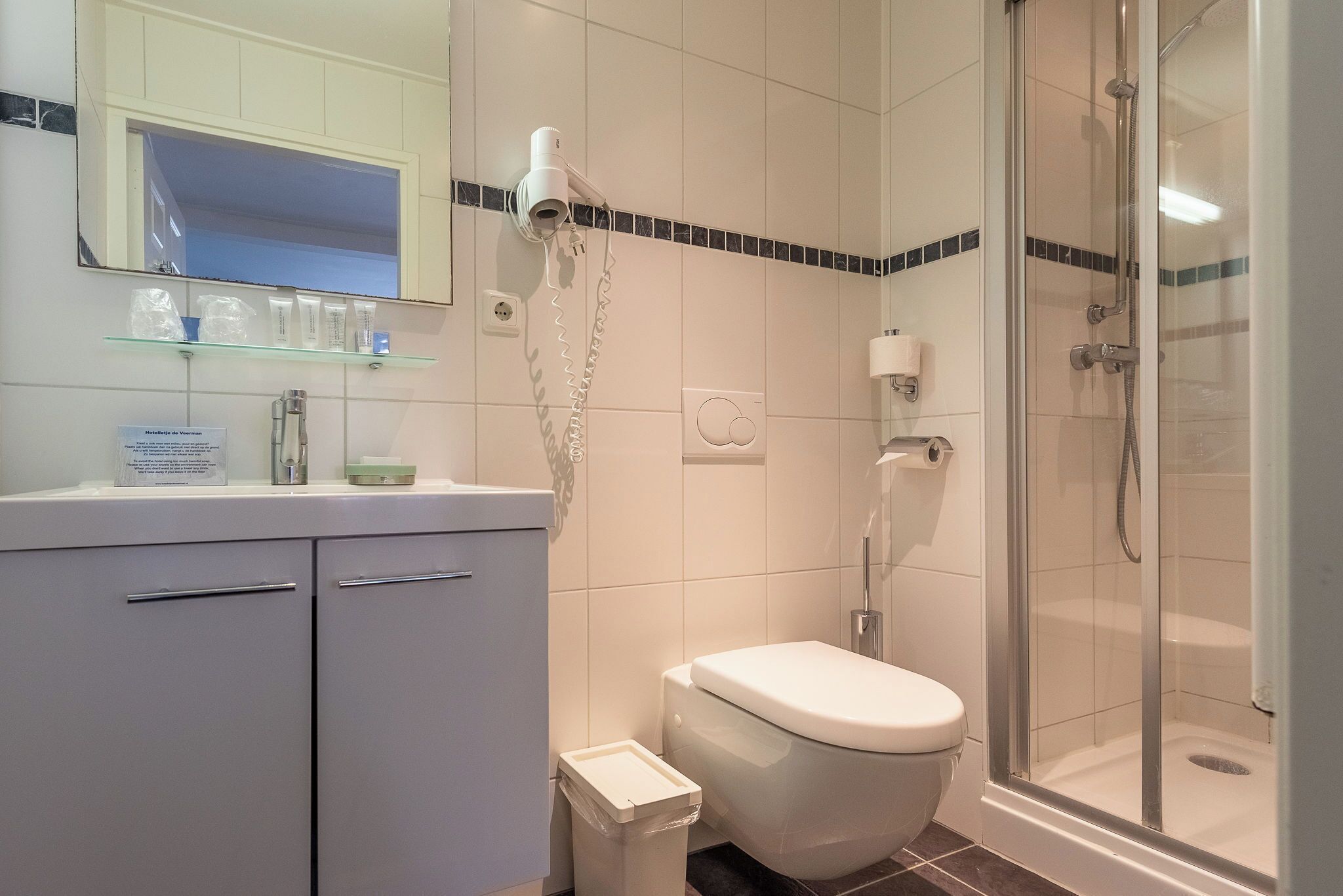 A bathroom with a toilet, a glass shower enclosure, a vanity with a mirror, and a hairdryer mounted on the wall.
