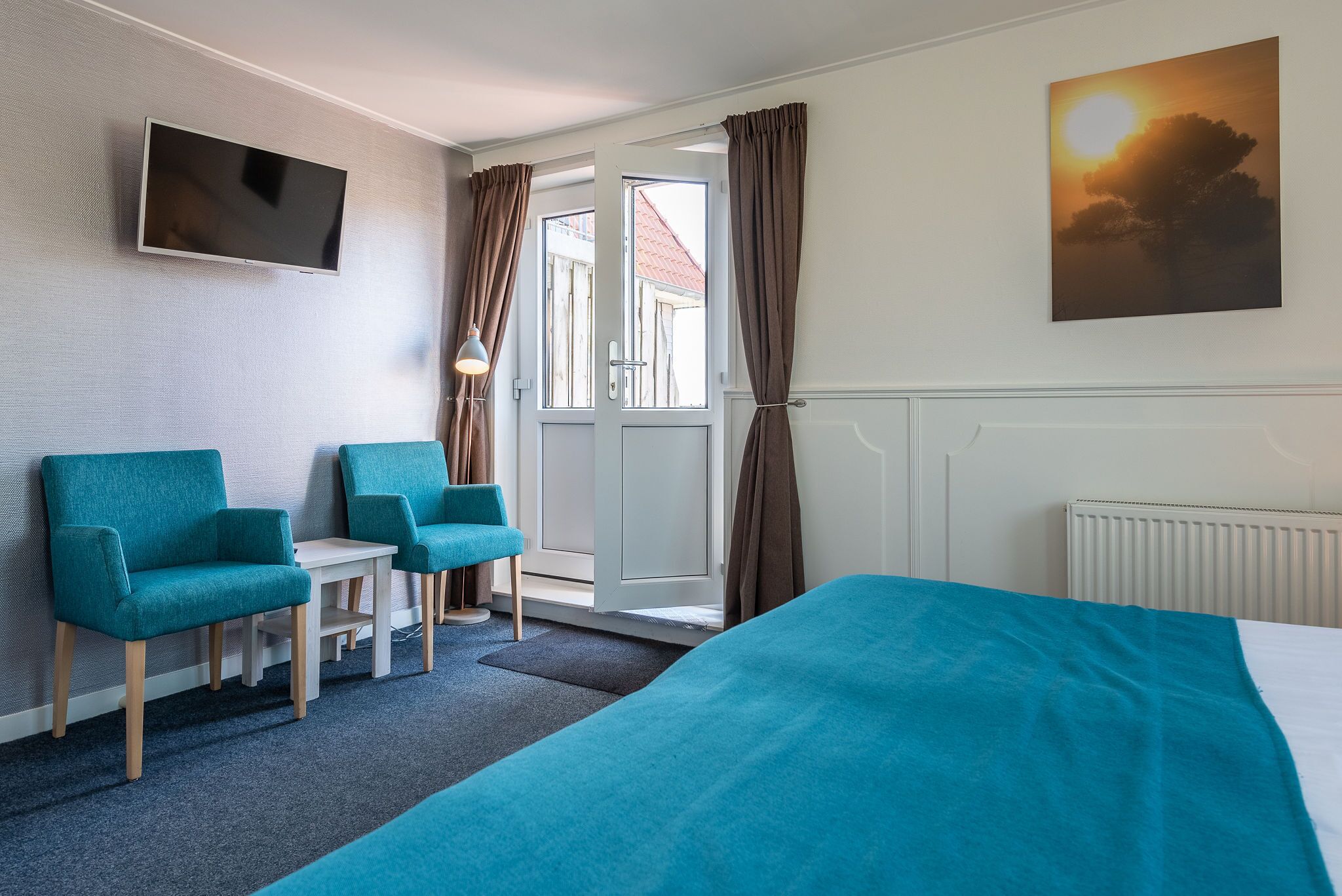 A hotel room with a bed, two teal armchairs, a small table, a TV mounted on the wall, and a painting of a sunset.