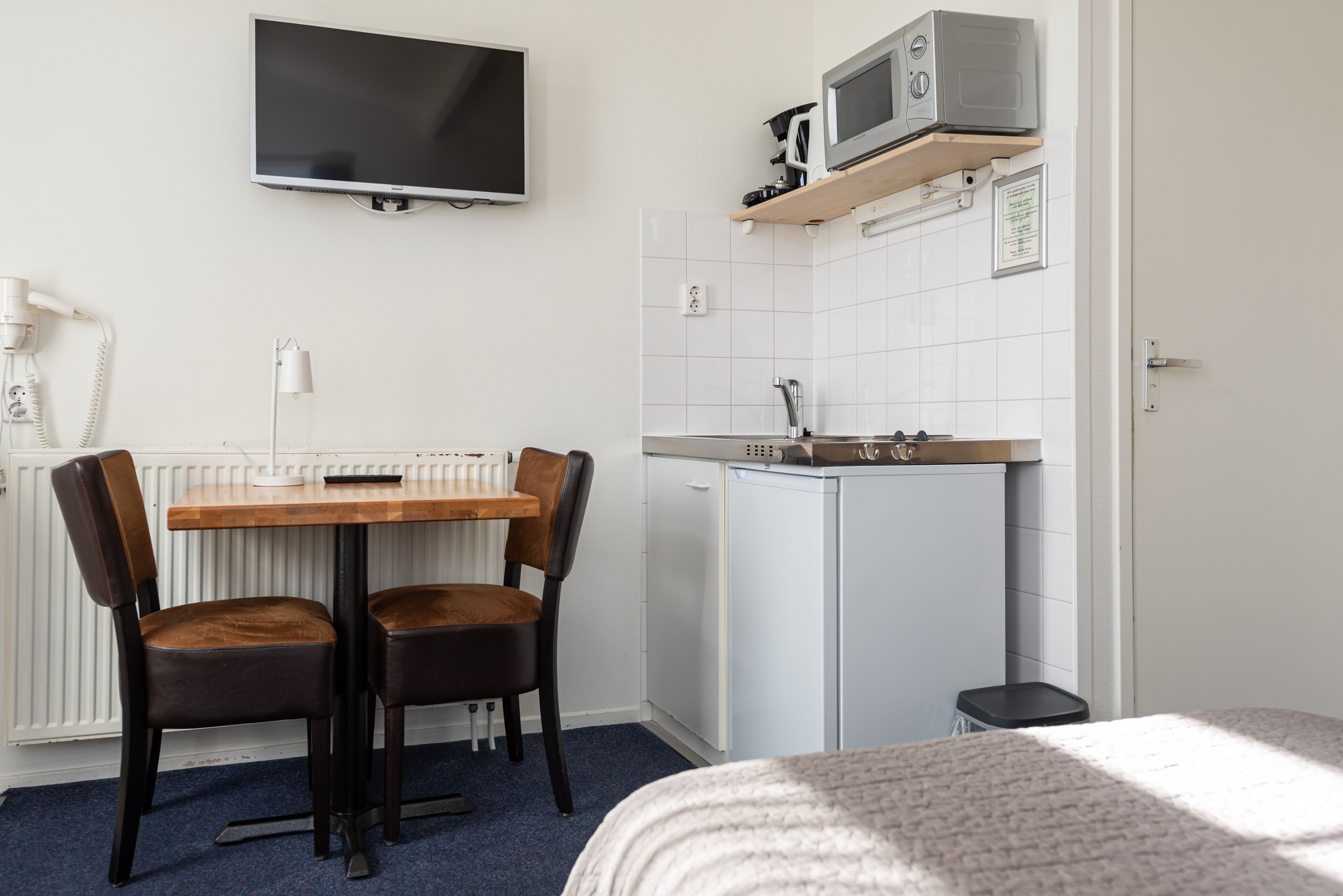 A compact room with a bed, a small kitchenette with a microwave, a sink, and a toilet. There is a table with two chairs and a TV mounted on the wall.