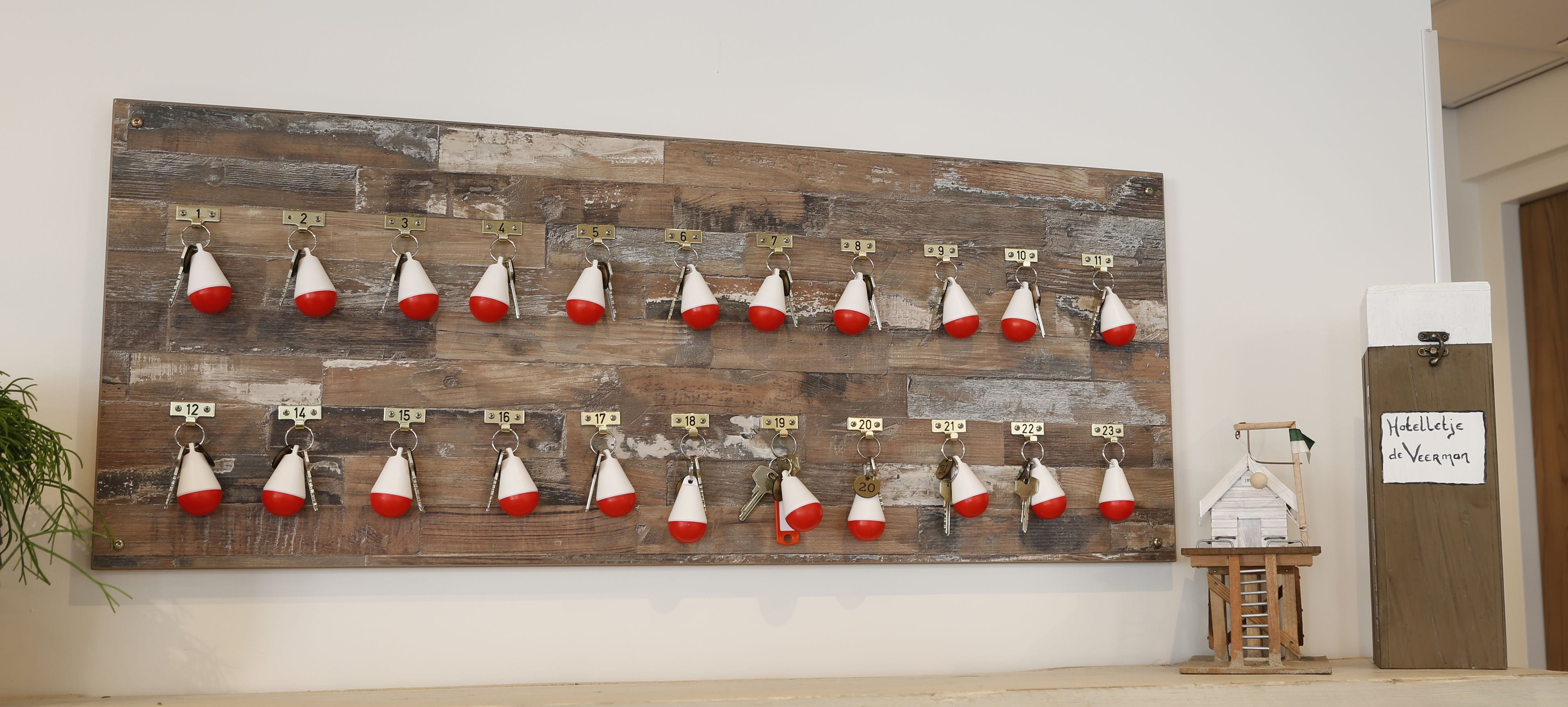 A wall-mounted key rack with numbered hooks, a small wooden model, and a sign reading "Hotelje de Verman".