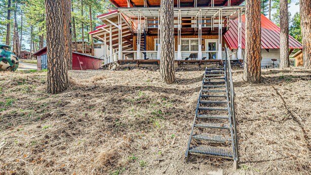 Una cabaña con terraza de madera, escalera metálica y techo rojo, rodeada de altos pinos.