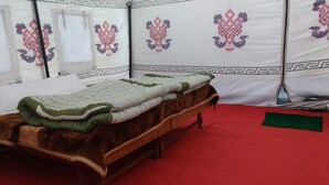 A bed with a green and white blanket, a red carpet, and a tent-like structure with decorative patterns in the background. - Jispa Journeys - Campsite (Lahul)