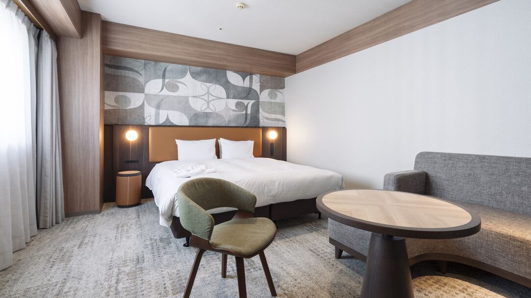 Una habitación de hotel con cama, una silla, un sofá, una mesita y un cabecero con diseño estampado.