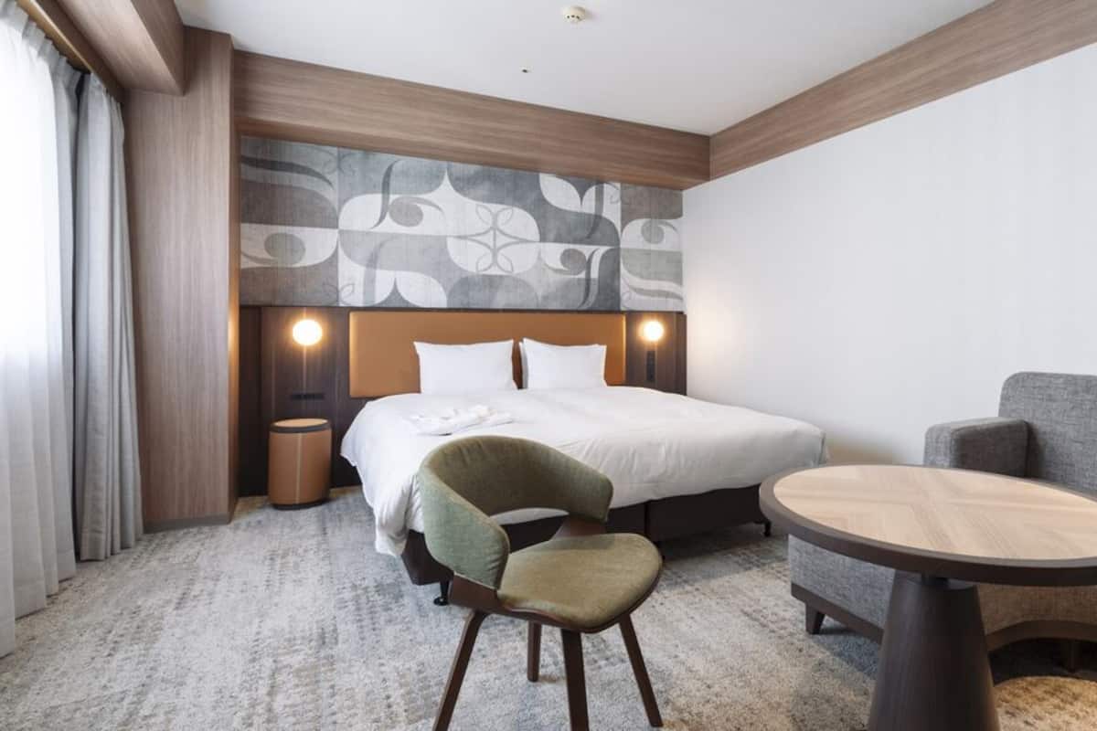 Una habitación de hotel con cama, una silla, un sofá, una mesita y un cabecero con diseño estampado.