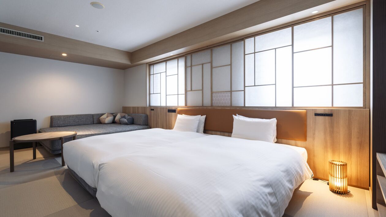 A modern hotel room with a large bed, a sofa, a bedside table, and a wall with a traditional Japanese screen.