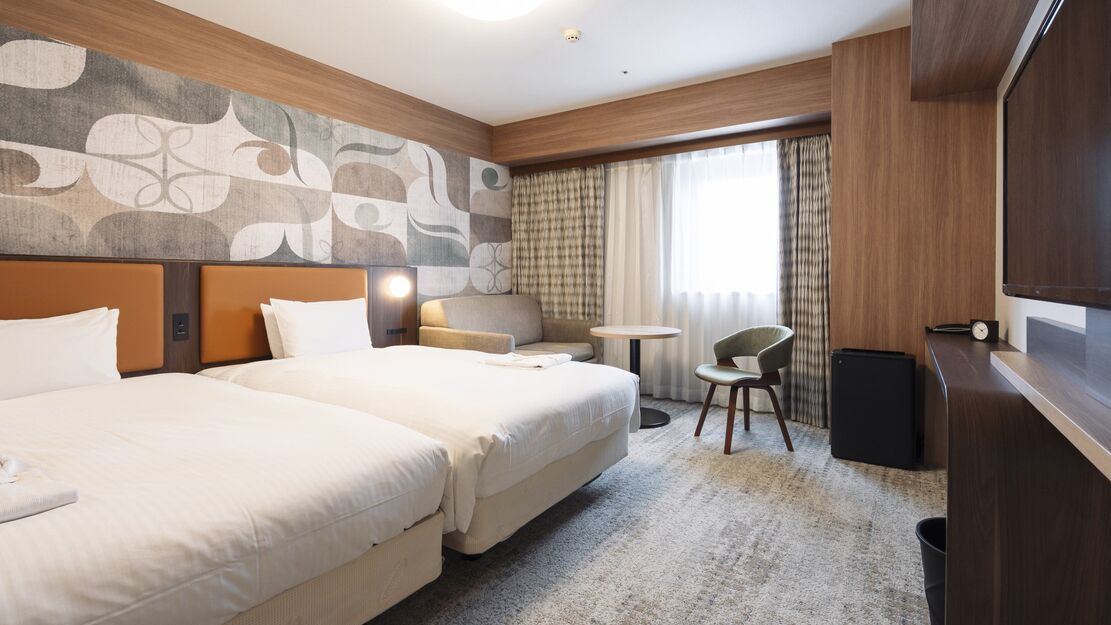 Habitación de hotel con dos camas, un sofá, una mesita, una silla, televisión y un ventanal con cortinas.