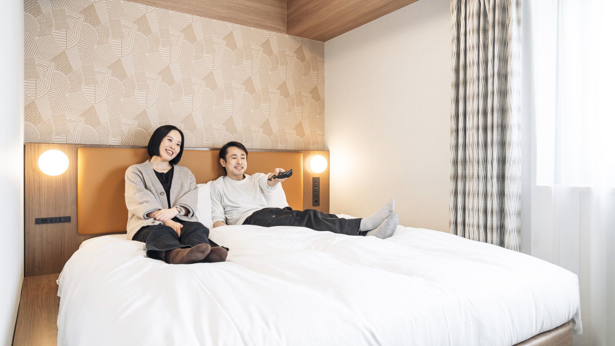 A hotel room with a large bed, two people sitting on it, a patterned wall, and a window with curtains.