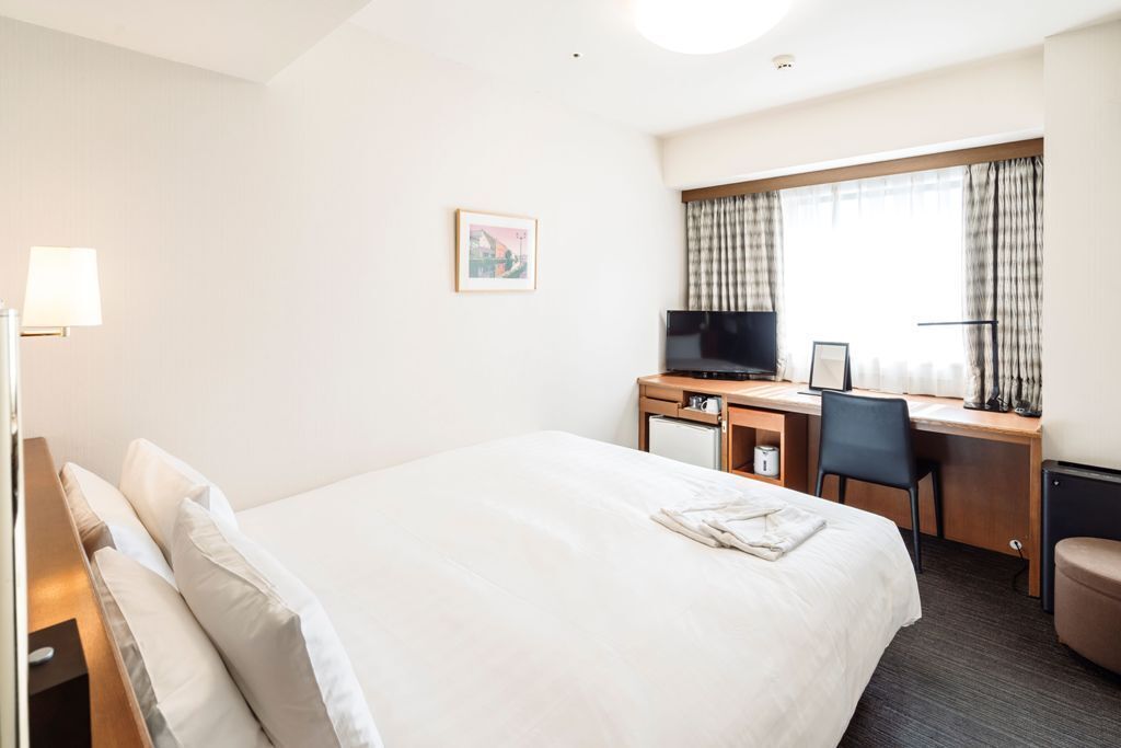 Habitación de hotel con una cama grande, un escritorio con computadora, televisión y ventana con cortinas.