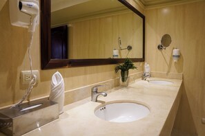 A hotel bathroom with two sinks, a large mirror, a hairdryer, and a towel dispenser.