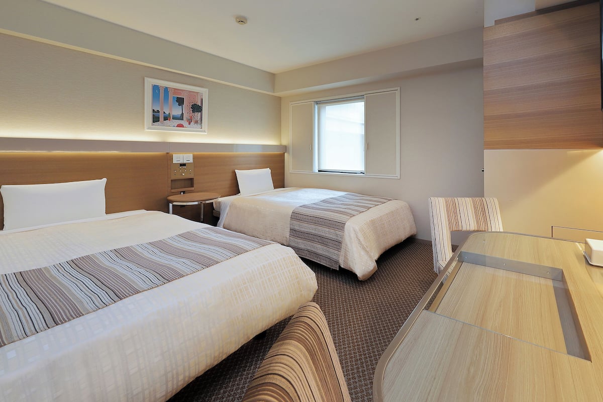 A hotel room with two beds, a desk, a chair, a window, and a painting on the wall.
