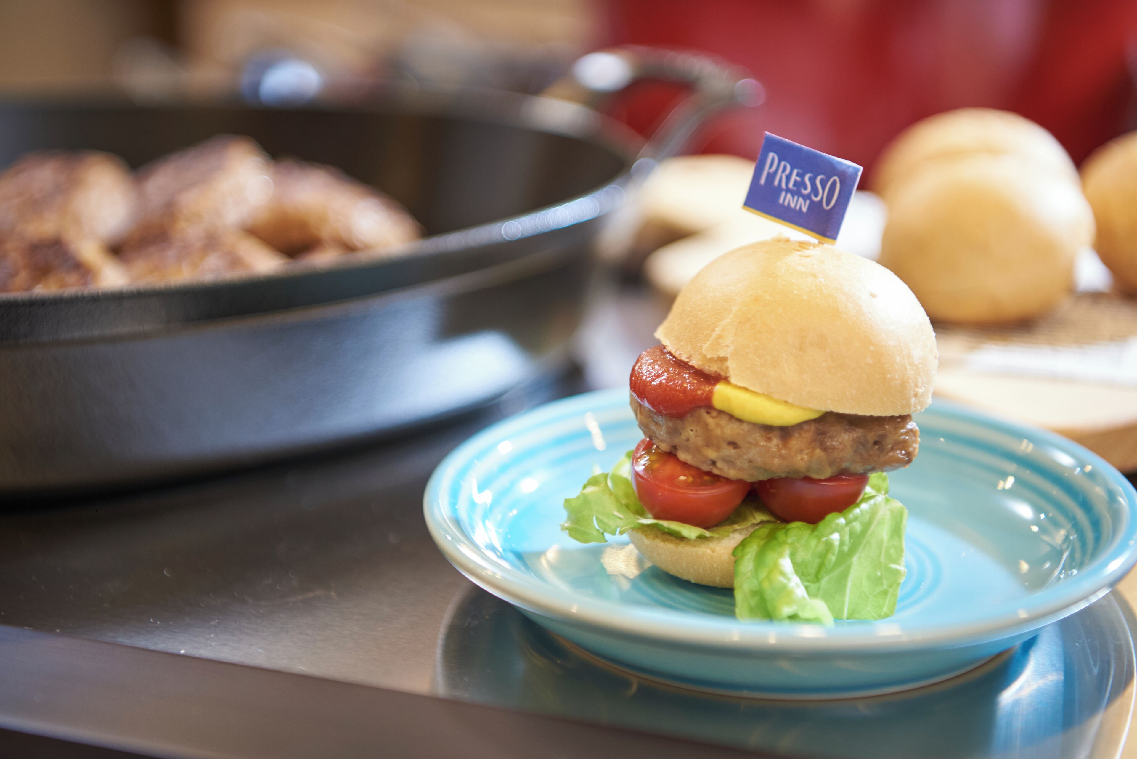 A sandwich on a blue plate with a 'PRESSO INN' sign, a pan of food in the background, and a bowl of onions.