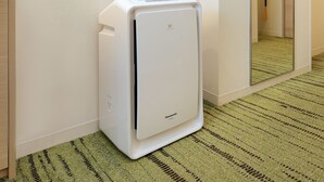 A white Panasonic air conditioner in a corner of a room with a green patterned carpet and a white textured wall. - Keio Presso Inn Gotanda (Tokyo)