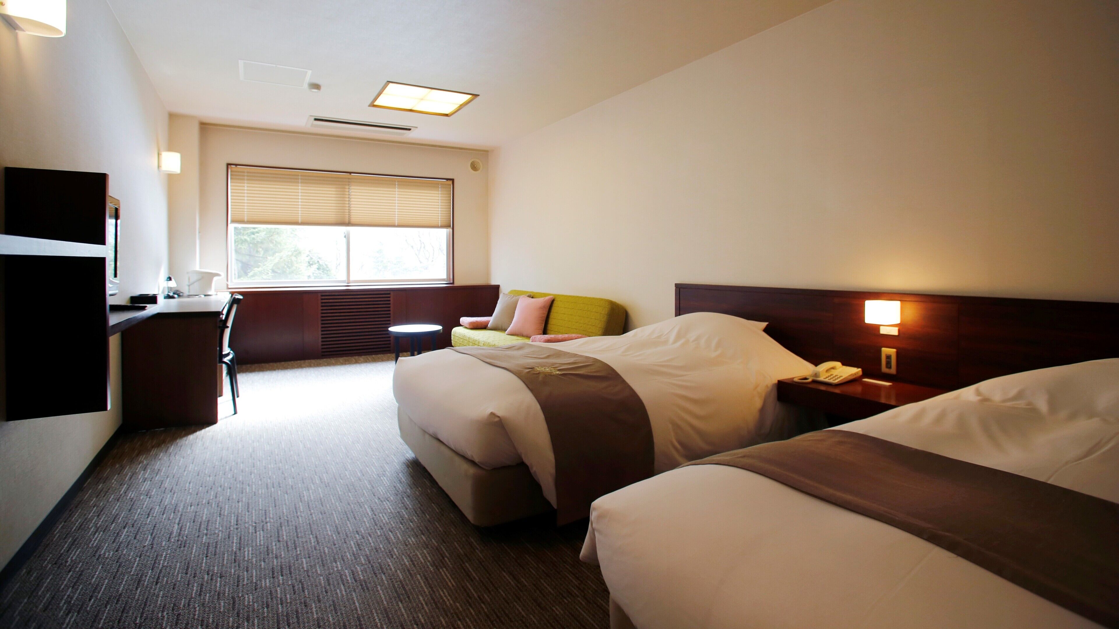Ein Hotelzimmer mit zwei Betten, einem Schreibtisch, einem Stuhl, einem Fenster mit Jalousien und einer Nachttischlampe.