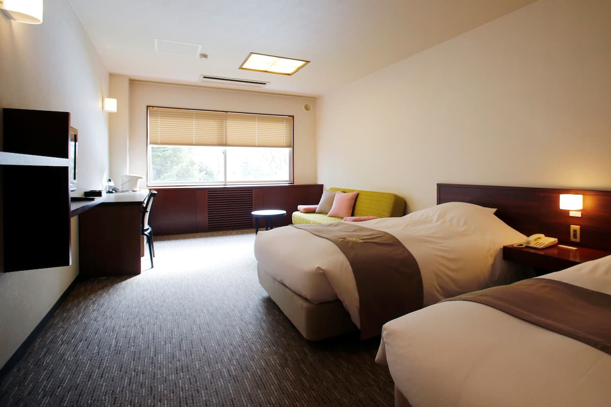 A hotel room with two beds, a desk, a chair, a window with blinds, and a bedside lamp.