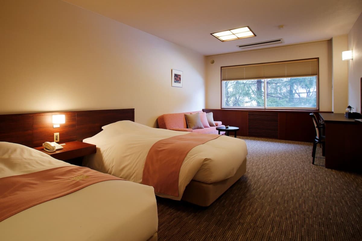 A hotel room with two beds, a desk, a chair, a window with a view, and a bedside lamp.