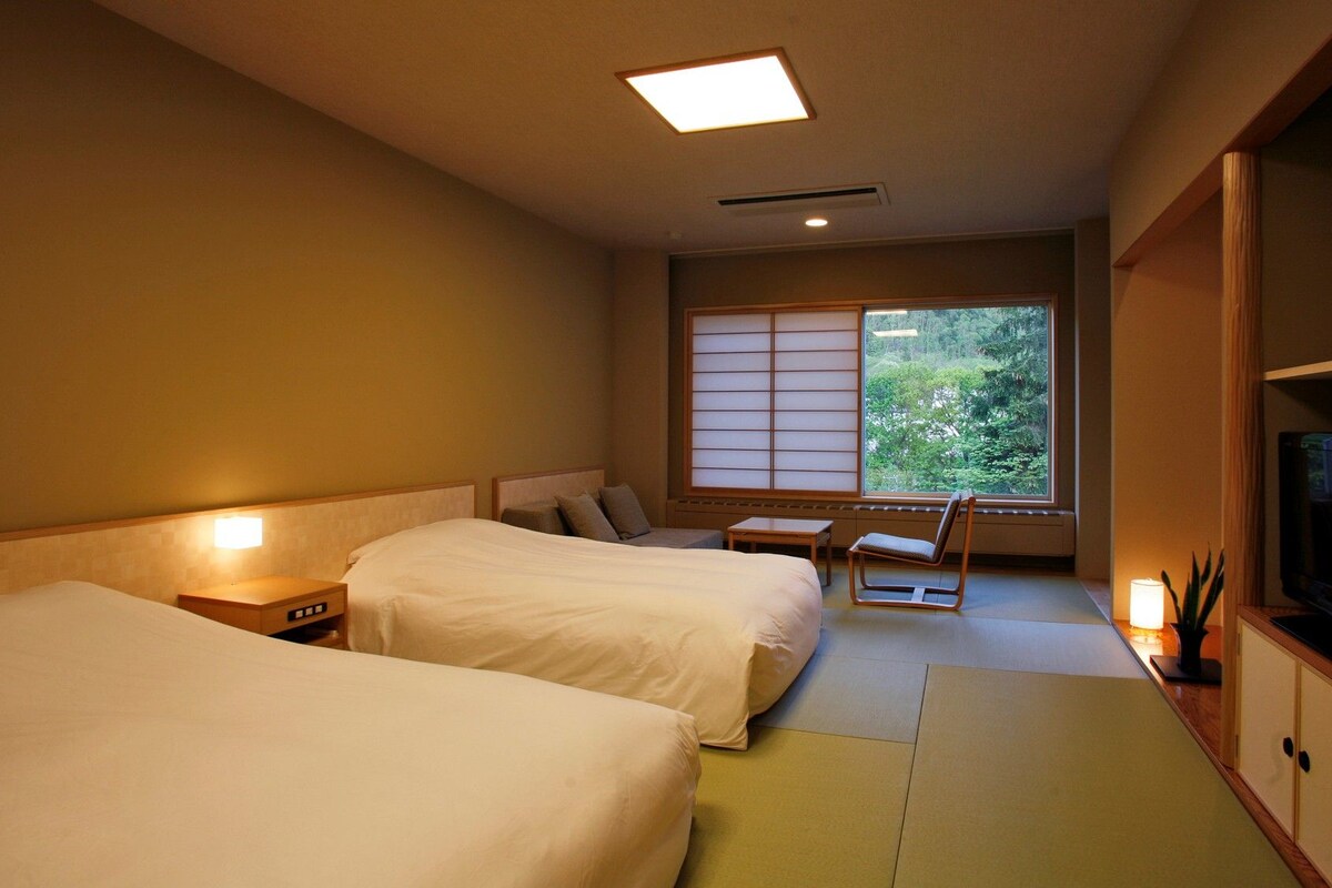 A hotel room with two beds, a small table, a chair, a TV, and a large window with a view of greenery.