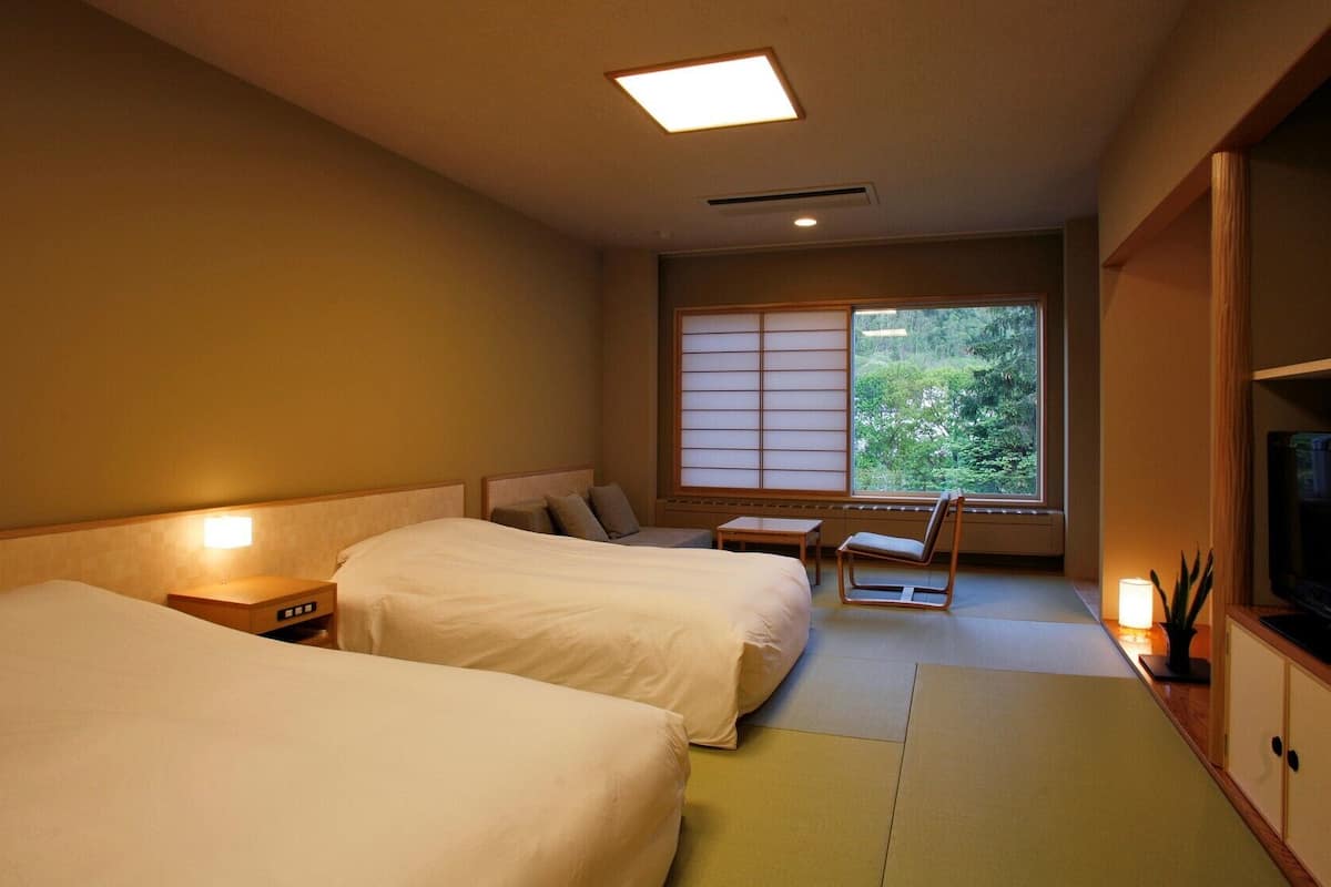 A hotel room with two beds, a small table, a chair, a TV, and a large window with a view of greenery.