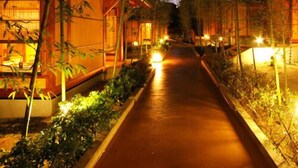 A pathway flanked by buildings with warm lighting, surrounded by trees and plants.