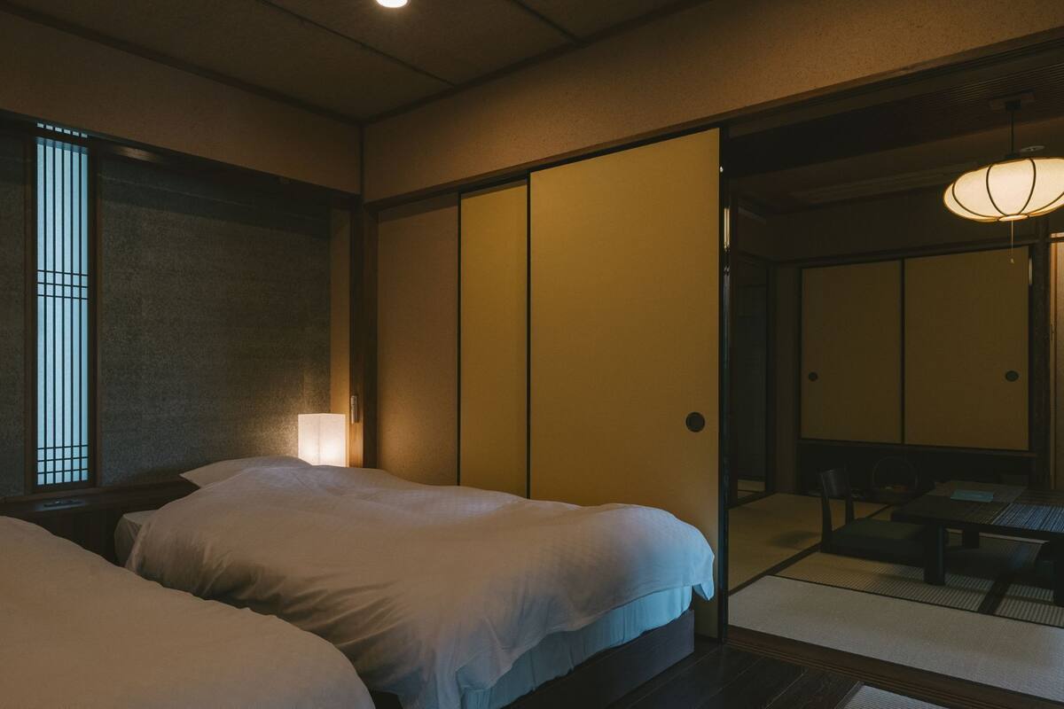 Ein traditioneller japanischer Raum mit zwei Betten, einer Schiebetür und einem kleinen Tisch mit Stühlen.