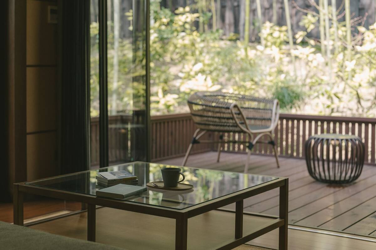 Ein modernes Zimmer mit Glastisch, einem Sessel und Blick auf eine Holzterrasse und grüne Vegetation.