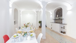 Comedor con una larga mesa dispuesta para una comida, paredes blancas y una cocina con salpicadero de azulejos.